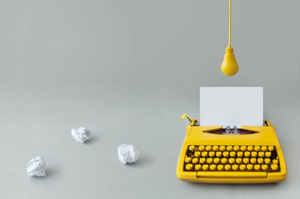 Yellow typewriter with plain white paper and a light bulb hoovering over it, and scrunched up paper on the floor beside it.