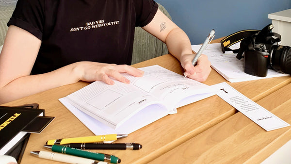Person sitting at a desk with papers and writing instruments, wearing a black t-shirt with text.