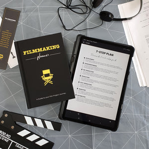 Filmmaking Planner Hardback, A5, yellow and black next to the digital planner on IPad with 7-step plan page displayed. 