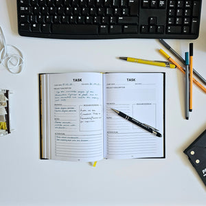 Open planner with task listings on a desk with a keyboard and pens.