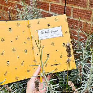 Yellow notebook with 'This book belongs to' tag held by a hand with plants in the background